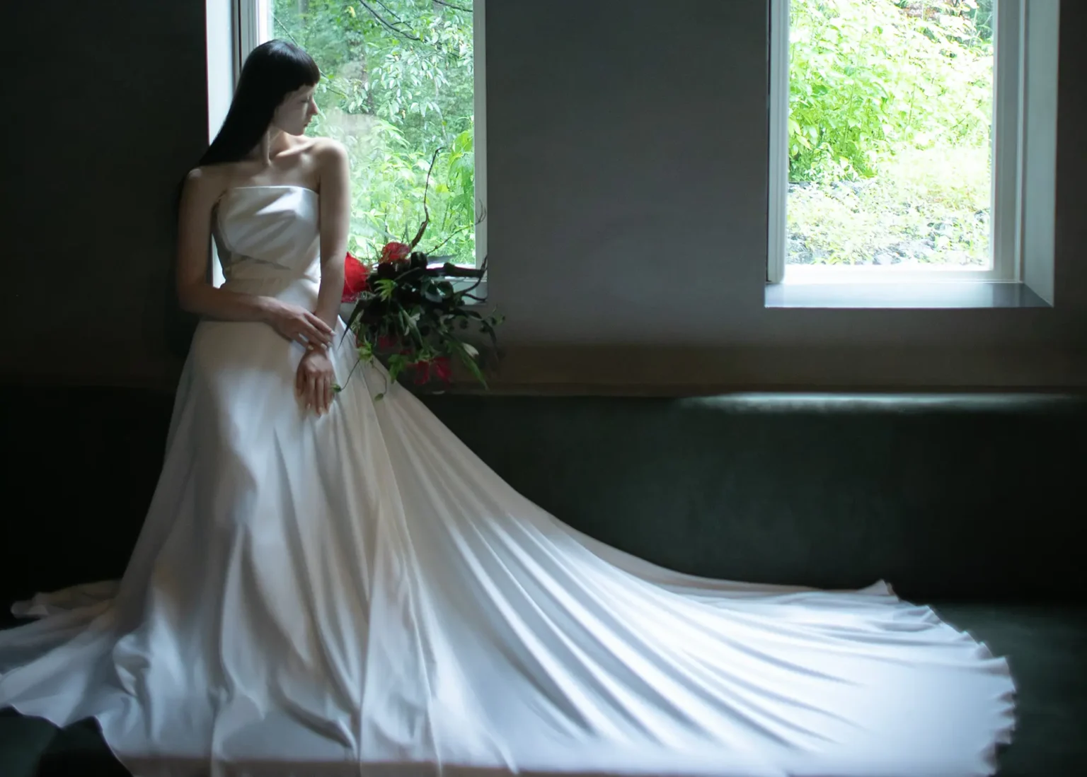 スレンダーラインのウェディングドレスを着て赤い花のブーケを持ち窓辺に座る女性